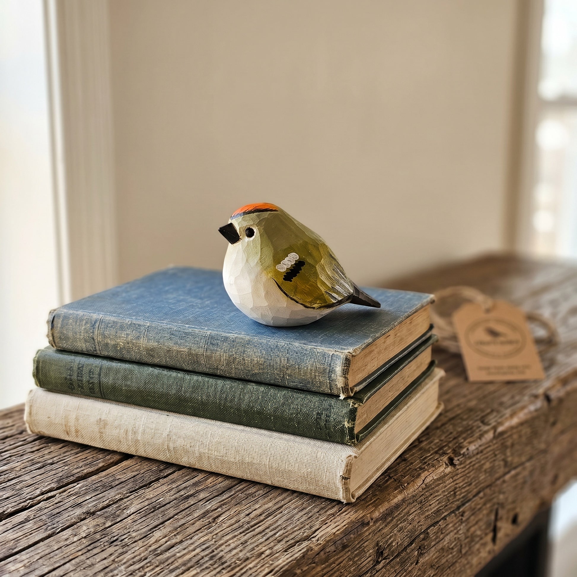 A small sculpted Golden-crowned kinglet figurine with olive green, white, black, and orange details sits on top of a stack of three vintage books on a rustic wooden table. A small paper tag is attached near the bird.