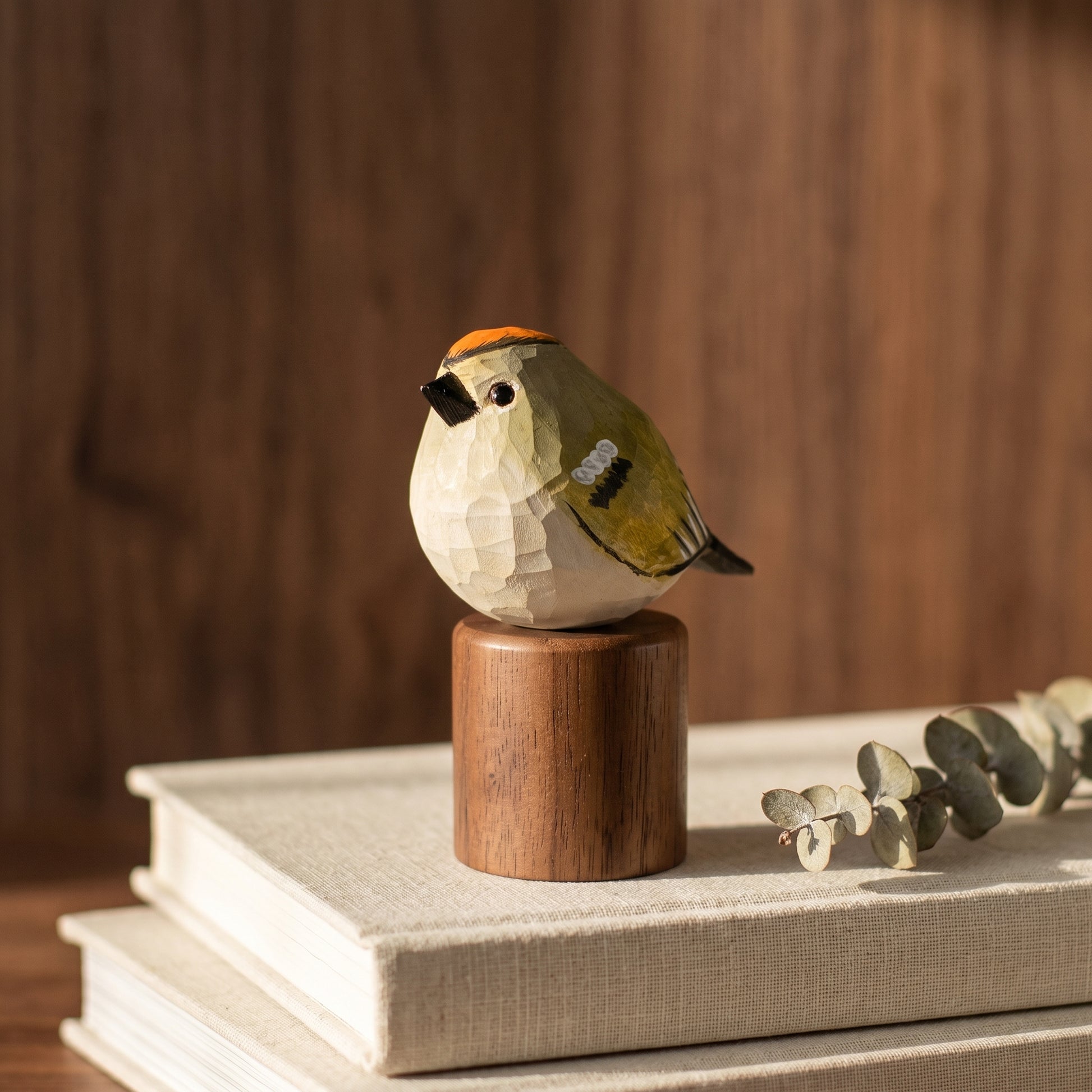 A small carved Golden-crowned kinglet figurine with a cream body, olive-green wing, black beak, and orange crown, perched on a cylindrical wooden base. The bird sculpture is styled on top of two stacked books with a sprig of eucalyptus against a warm wood background.