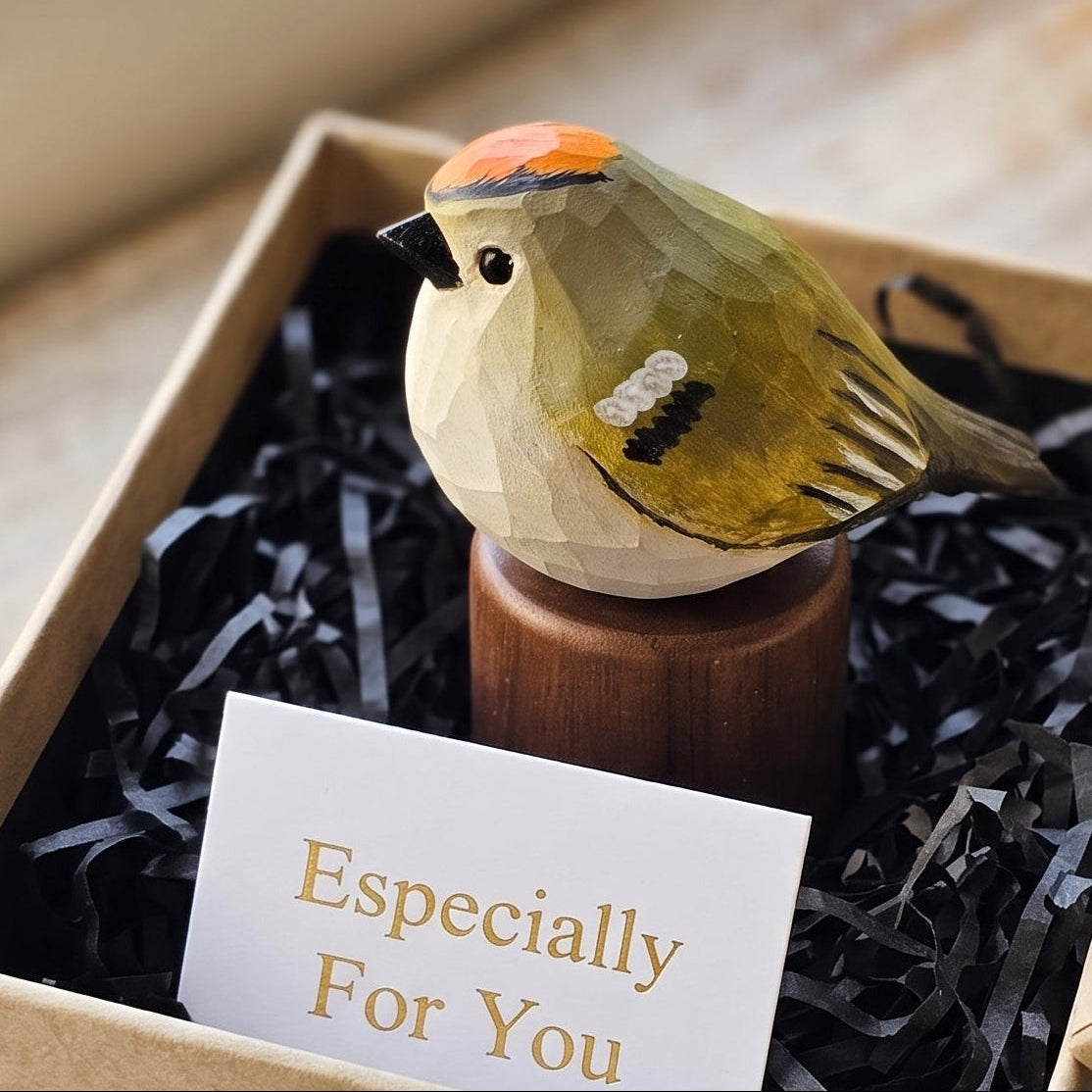 A small painted Golden-crowned kinglet figurine on a wooden stand presented inside a kraft gift box with black shredded paper filler and a card that reads 'Especially For You.' The box is open on a rustic wooden surface near a window.