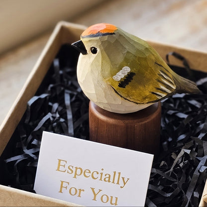 A small painted Golden-crowned kinglet figurine on a wooden stand presented inside a kraft gift box with black shredded paper filler and a card that reads 'Especially For You.' The box is open on a rustic wooden surface near a window.