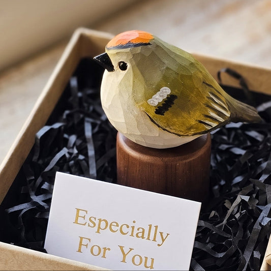 A small painted Golden-crowned kinglet figurine on a wooden stand presented inside a kraft gift box with black shredded paper filler and a card that reads 'Especially For You.' The box is open on a rustic wooden surface near a window.