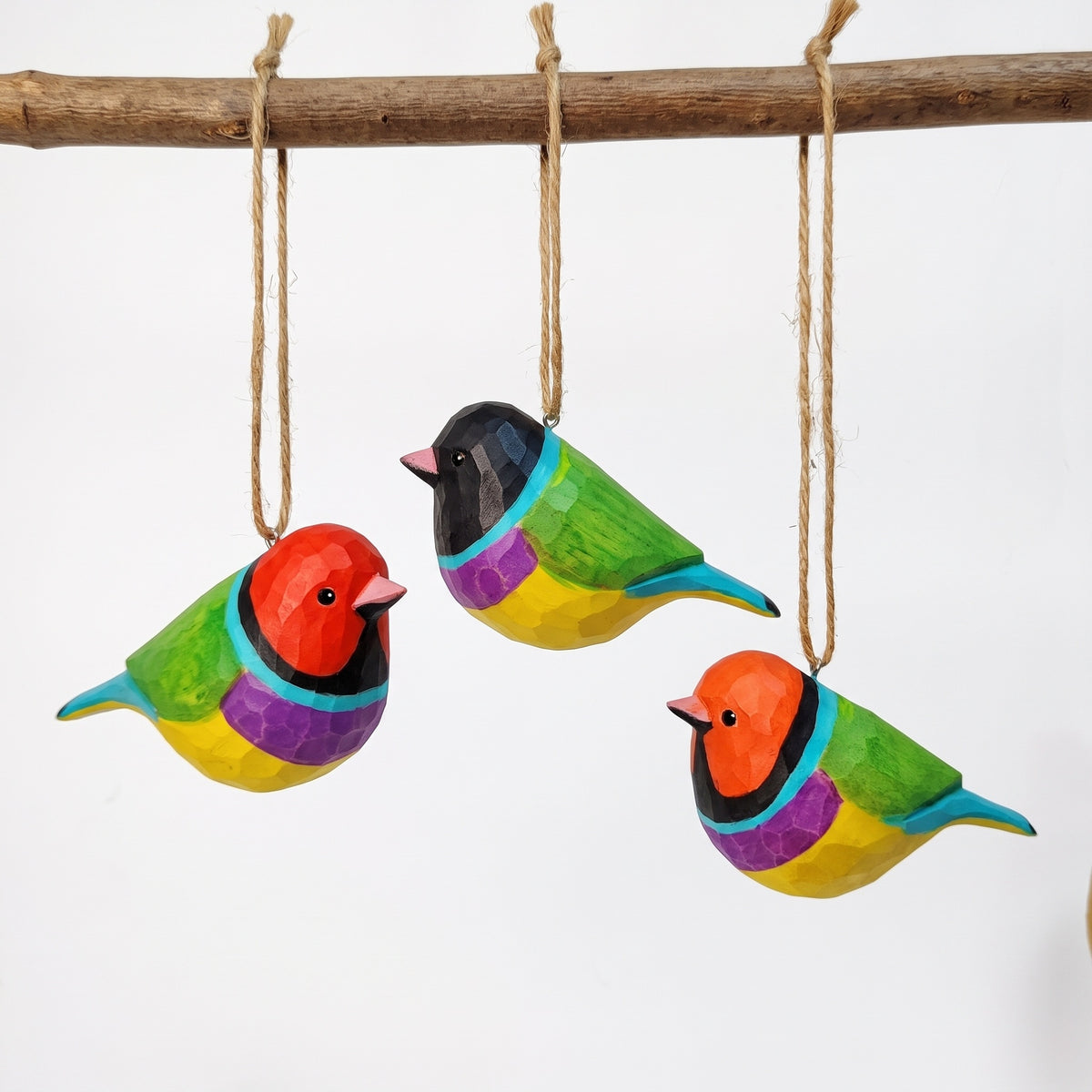 Three colorful wooden Gouldian finch ornaments hanging from a natural branch with jute ropes against a plain white background.
