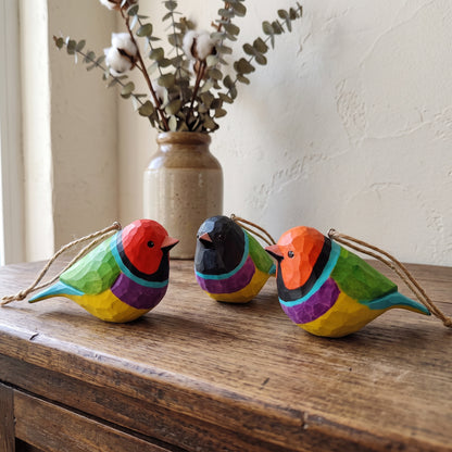Three colorful wooden Gouldian finch ornaments with geometric facets and jute hanging strings, displayed on a rustic wooden table with a vase of cotton stems in the background.