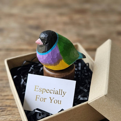 A handcrafted wooden Gouldian finch figurine with vibrant painted colors sits in an open kraft gift box filled with black paper shred and a small card that reads 'Especially For You'. The box is placed on a rustic wooden surface.