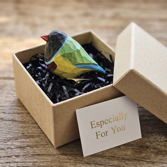 A close-up side-profile shot of a hand-carved wooden Orange-headed Gouldian Finch clip-on ornament resting on a plain white surface, featuring an orange-red face, a dark eye, a bright turquoise-blue nape collar, a grass-green back, a purple-violet breast, a bold yellow belly, light blue wing tip details, and a silver metal clip attached underneath.