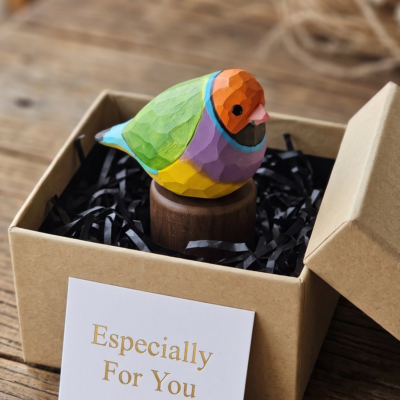 A small colorful geometric Gouldian finch figurine displayed inside an open kraft gift box with black crinkle paper filler and a card that reads 'Especially For You,' set on a rustic wooden table.