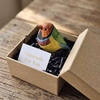 A hand-carved wooden Orange-headed Gouldian Finch clip-on ornament resting inside an open kraft gift box with black shredded paper and a white "Especially For You" card, set on a rustic wooden surface.