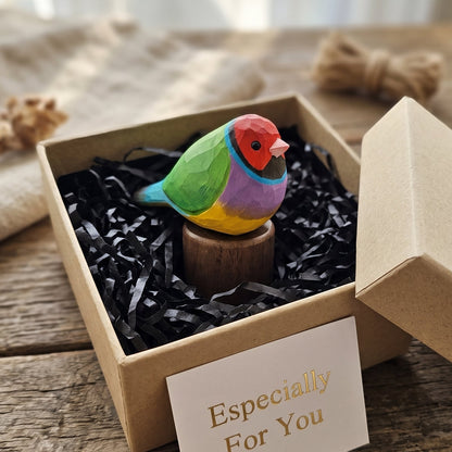 A small hand-painted Gouldian finch figurine with vibrant green, blue, yellow, purple, and red feathers sits in a brown kraft gift box filled with black shredded paper. A white card reading 'Especially For You' is placed in front, and the box lid leans against the right side on a rustic wooden surface.