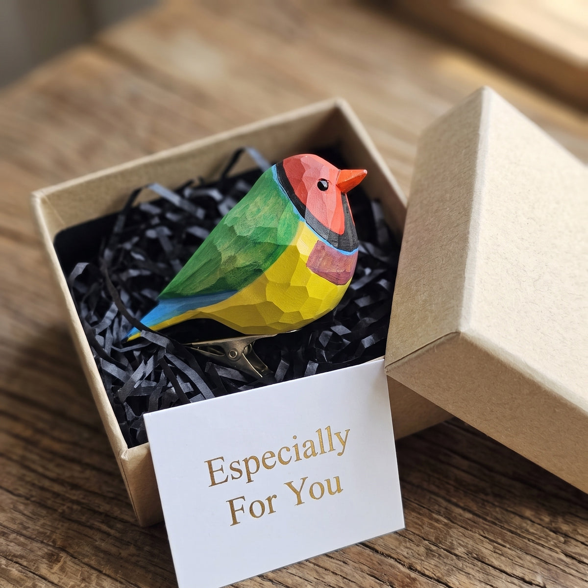 A hand-carved wooden Red-headed Gouldian Finch clip-on ornament resting inside an open kraft paper gift box filled with black shredded paper, featuring a red face, a dark eye, a turquoise-blue nape collar, a grass-green back, a purple-violet breast, a bold yellow belly, and a white card reading "Especially For You" placed in the foreground on a rustic wooden surface.
