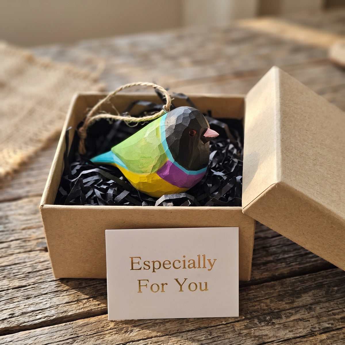 A colorful faceted Gouldian finch ornament nestled in black crinkle paper inside a kraft gift box, with a small card reading 'Especially For You' in front. The bird is decorated with bright green, yellow, purple, blue, and black sections, and the box sits on a rustic wooden surface.