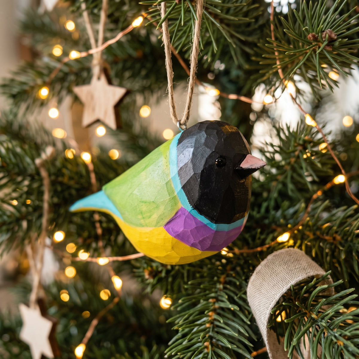 A colorful wooden Gouldian finch ornament hanging on a decorated Christmas tree with warm string lights. The ornament has a faceted geometric design with green, yellow, purple, and black sections, and a small pink beak.