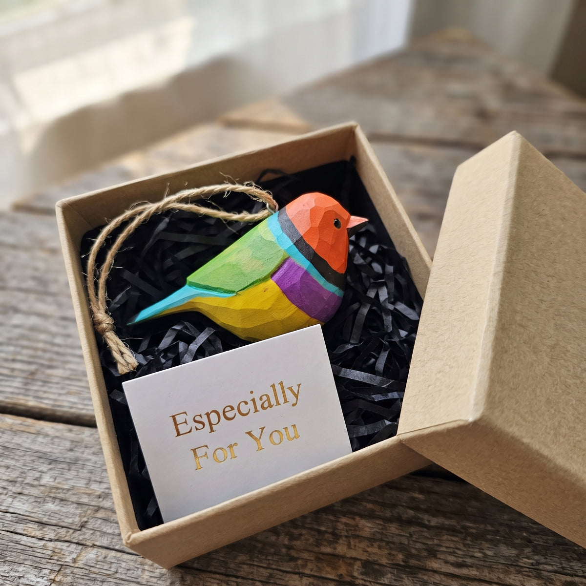 A colorful wooden Gouldian finch ornament with a string loop, presented in a kraft gift box with black shredded paper and a card that reads 'Especially For You'.