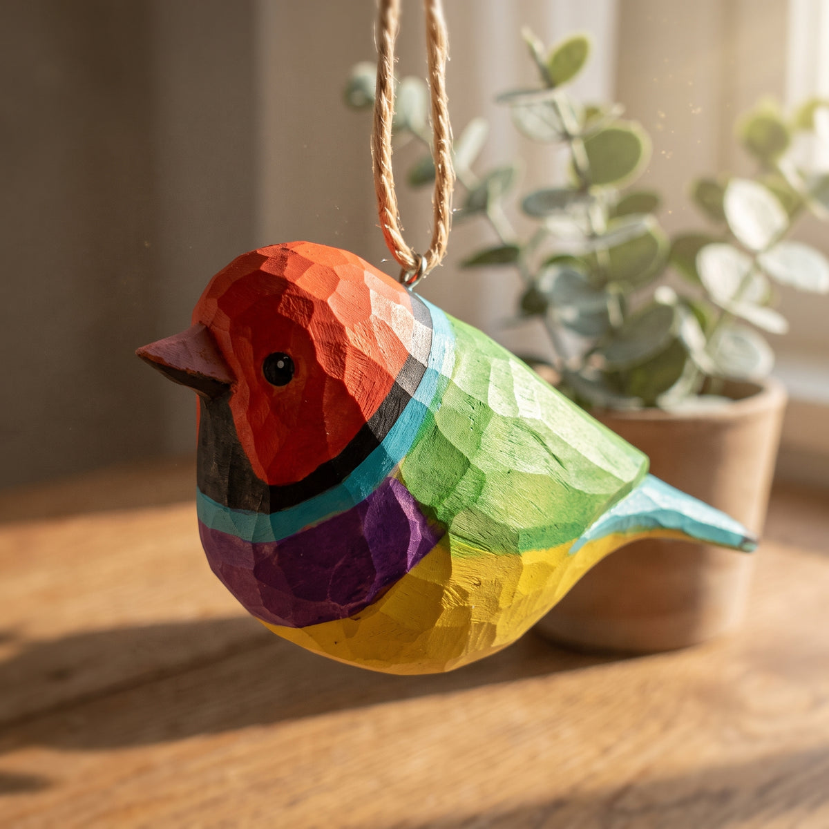 A colorful wooden Gouldian finch ornament with a geometric, faceted carving style, hanging by a natural jute string. The bird features painted sections of red, orange, black, turquoise, green, purple, and yellow, set against a warm, softly blurred background with a potted eucalyptus plant.
