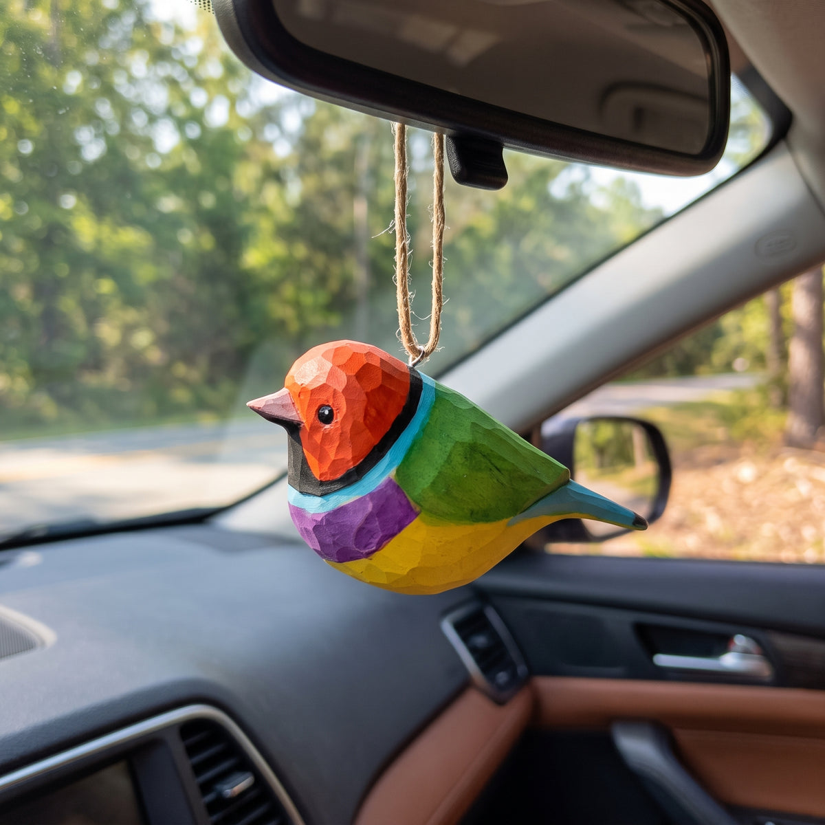 A colorful Gouldian finch-shaped car air freshener ornament hanging from a car's rearview mirror. The ornament features a geometric, faceted design with orange, green, yellow, purple, and blue sections, set against a blurred background of a car interior and trees.
