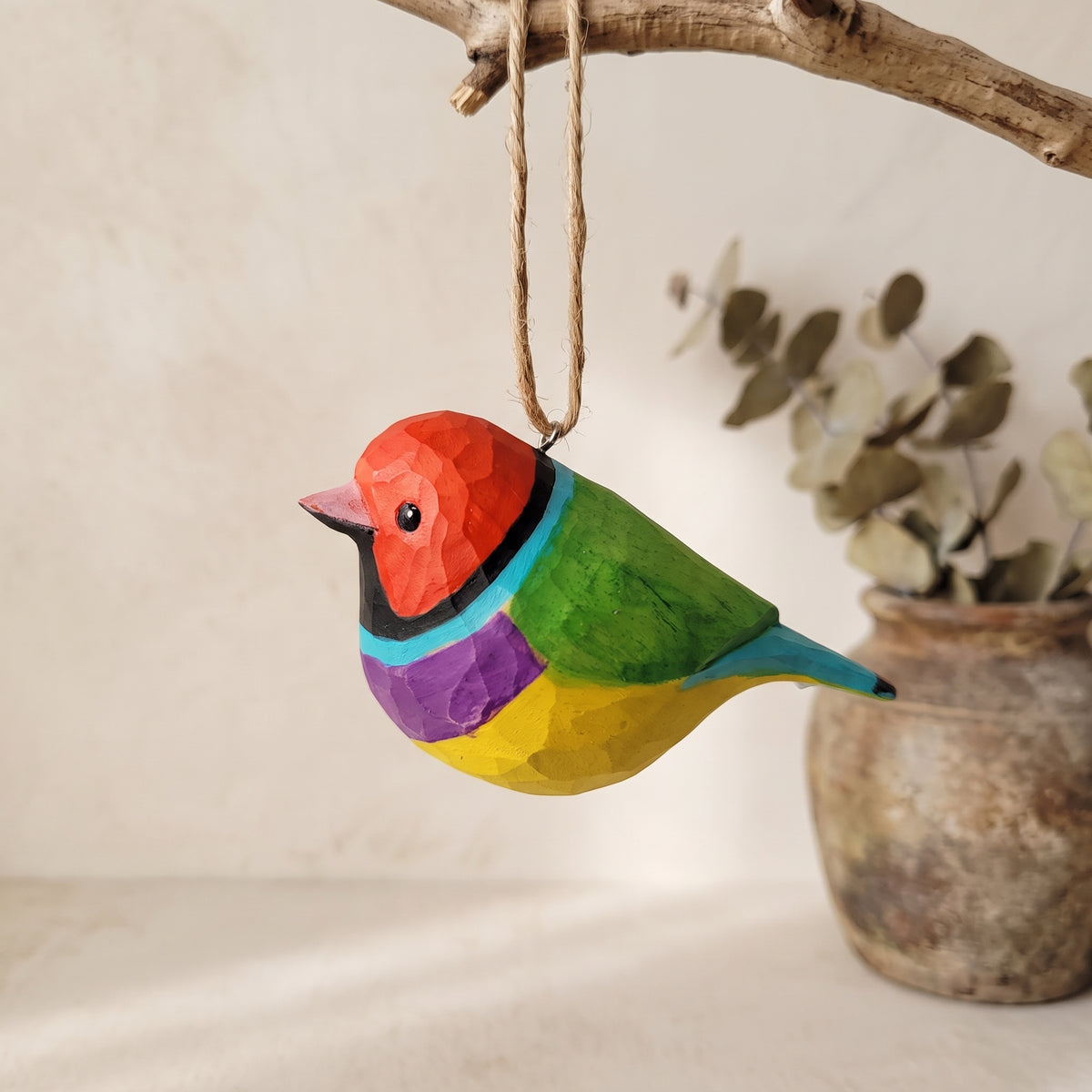 A colorful wooden Gouldian finch ornament hanging from a natural branch with twine, set against a softly blurred neutral background with a rustic pot.