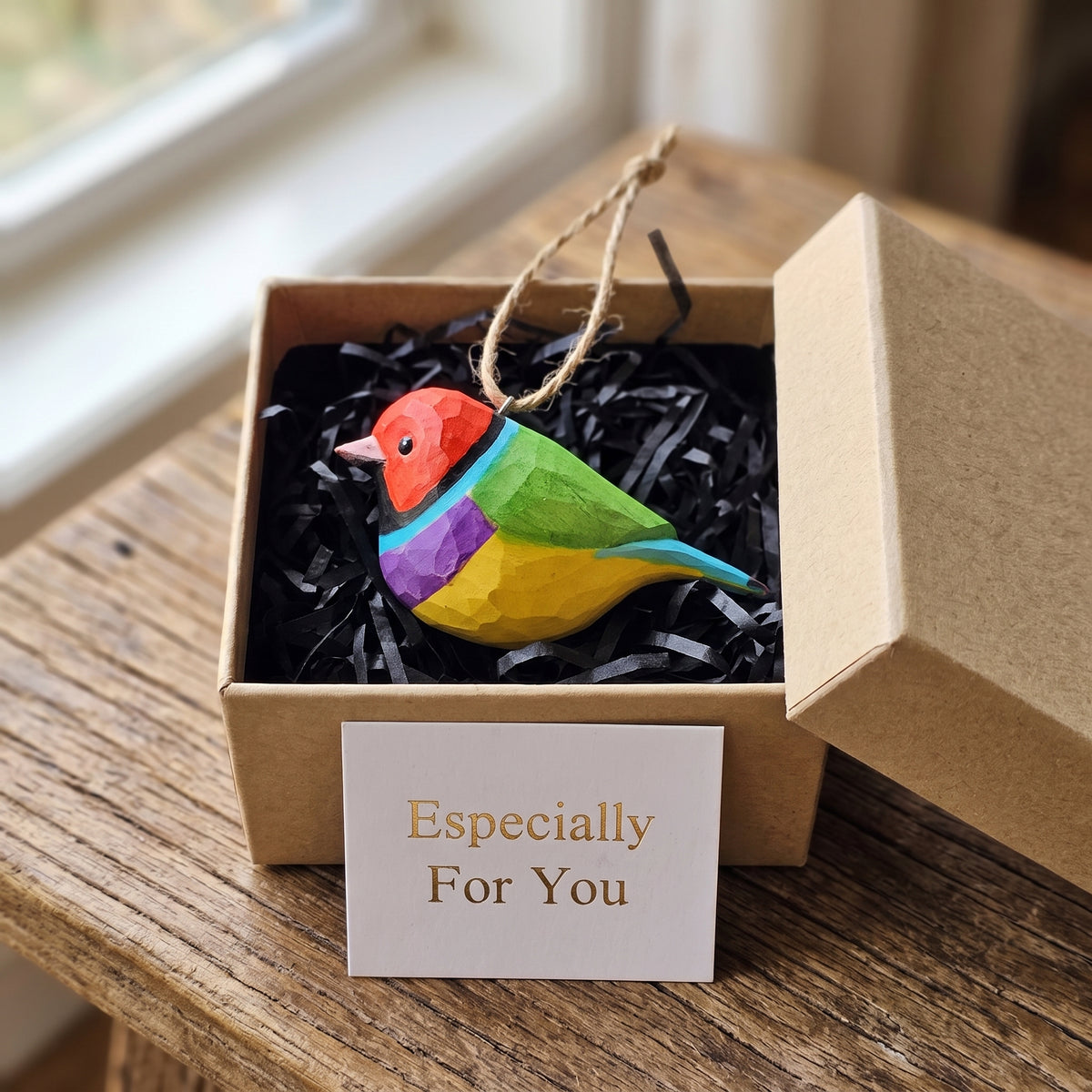 A colorful wooden Gouldian finch ornament with a red, yellow, green, and purple body sits in a brown gift box filled with black shredded paper. A small card in front reads 'Especially For You'.