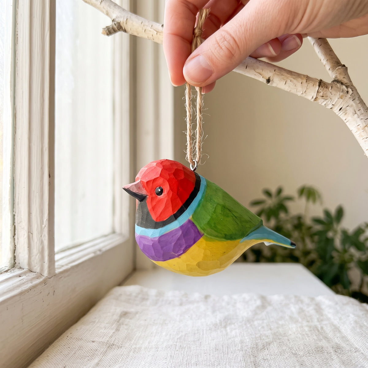 A colorful, low-poly Gouldian finch ornament with a red head, green back, purple and yellow underbelly, and blue tail, hanging from a jute loop on a birch branch near a window.