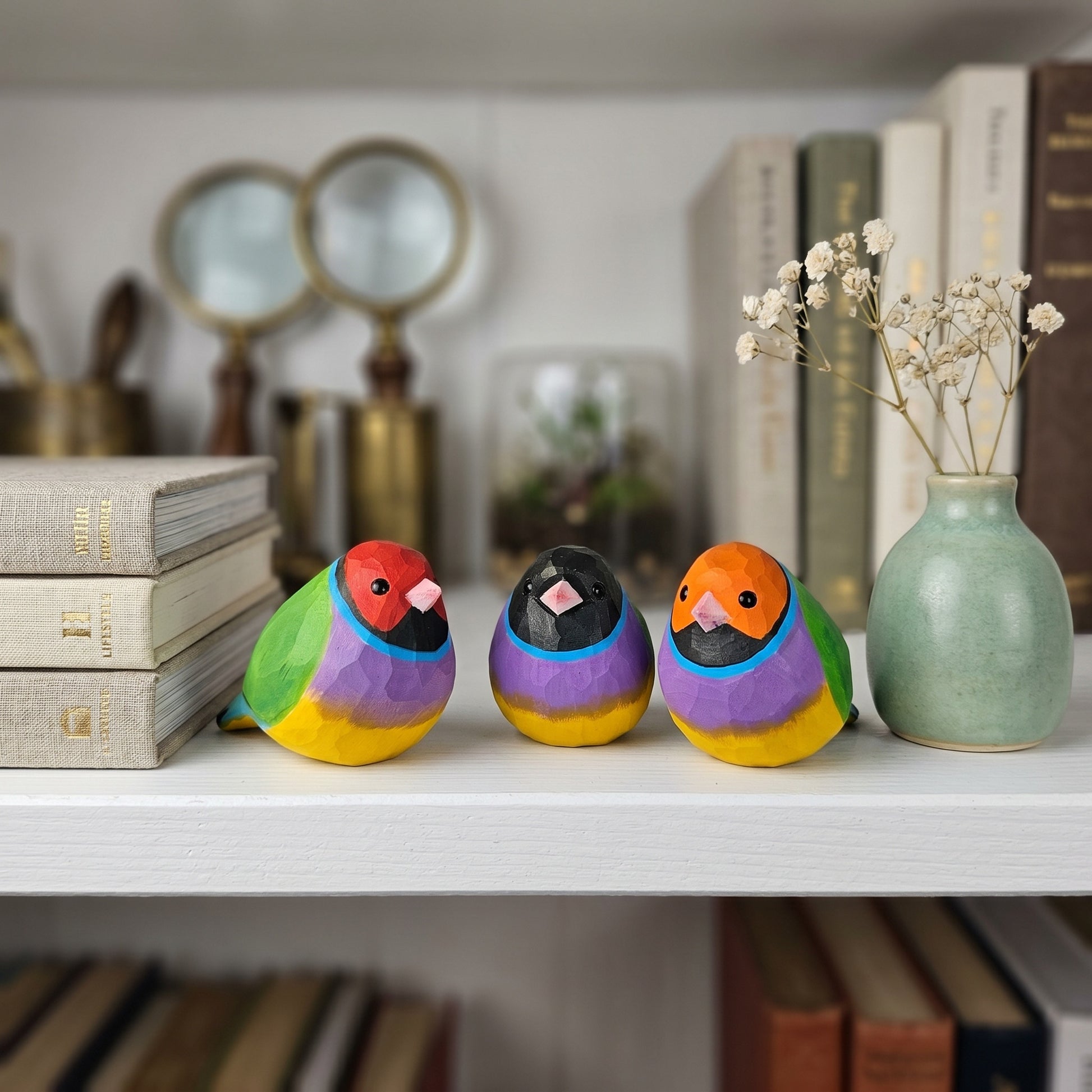 Three small hand-painted wooden Gouldian finch figurines in vibrant rainbow colors are arranged on a white shelf next to a light green ceramic vase with dried white flowers, with books and magnifying glasses blurred in the background.