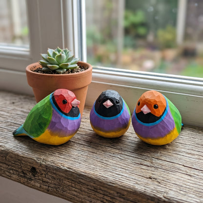 Three colorful, faceted Gouldian finch figurines painted like small birds, displayed on a rustic wooden windowsill next to a potted succulent.