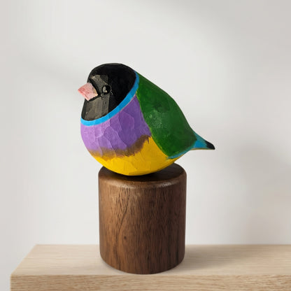 A handcrafted wooden Gouldian finch figurine with vibrant painted colors (black, pink, blue, purple, green, yellow) perched on a dark brown cylindrical wooden base, set against a plain light gray background.