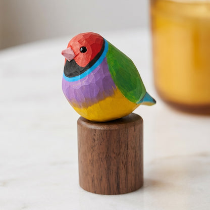 A small carved wooden Gouldian finch figurine with a colorful geometric pattern, perched on a cylindrical wooden base. The bird features a red head, black collar, blue band, purple chest, yellow belly, and green wing, set against a softly blurred background with a warm yellow candle.