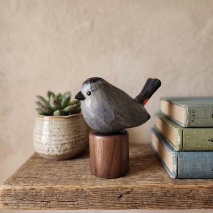 A carved wooden Gray Catbird figurine with a gray and black body, small orange accent on the tail, and a dark brown cylindrical base, displayed on a rustic wooden surface with a small potted succulent and stacked vintage books in the background.