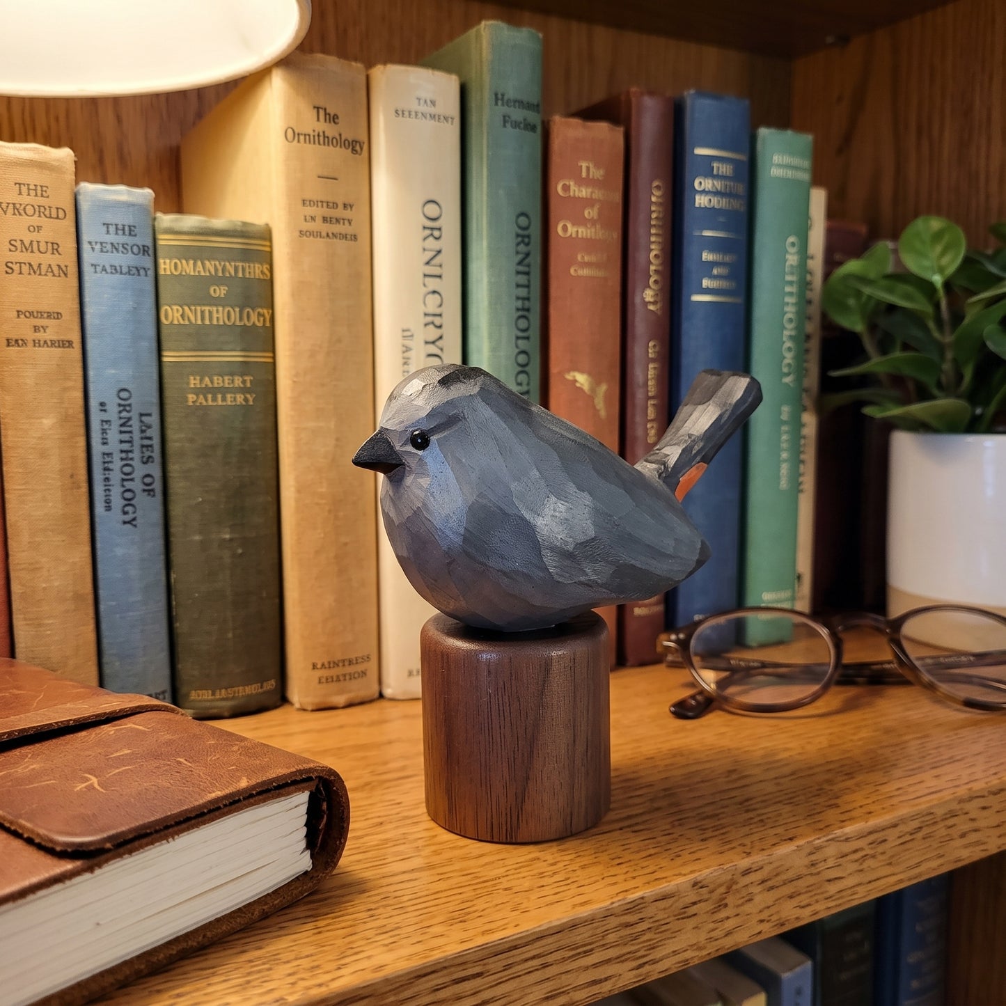 A carved wooden Gray Catbird figurine with a dark gray-blue body and a medium brown wooden base, displayed on a wooden bookshelf next to books, glasses, and a plant.