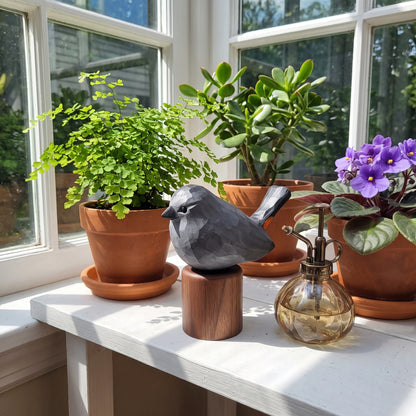 A gray Gray Catbird figurine perched on a wooden base sits on a white windowsill beside potted plants and a glass pump bottle in bright natural light.