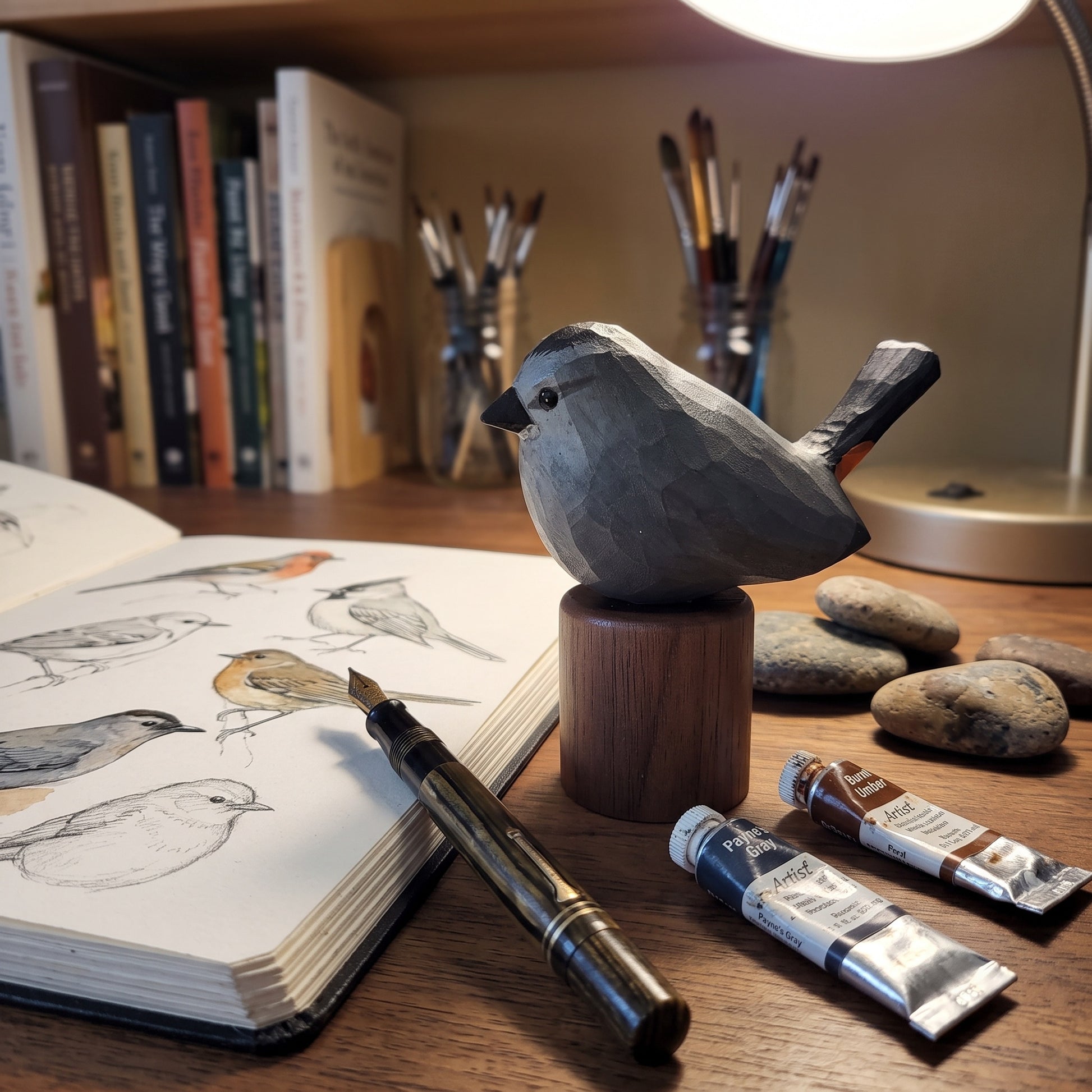 A carved wooden Gray Catbird sculpture on a wooden stand sits on a desk surrounded by art supplies, including an open sketchbook with bird drawings, a paintbrush holder, two paint tubes, and smooth stones. A lamp is visible in the background.
