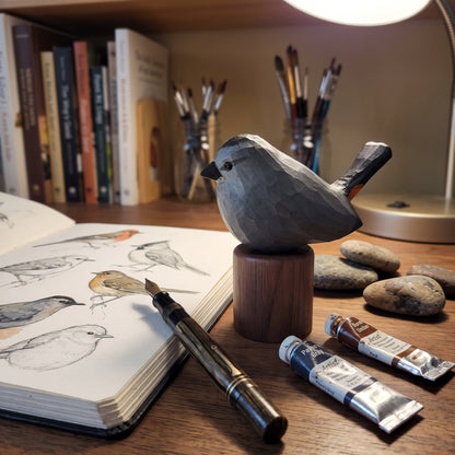 A carved wooden Gray Catbird sculpture on a wooden stand sits on a desk surrounded by art supplies, including an open sketchbook with bird drawings, a paintbrush holder, two paint tubes, and smooth stones. A lamp is visible in the background.