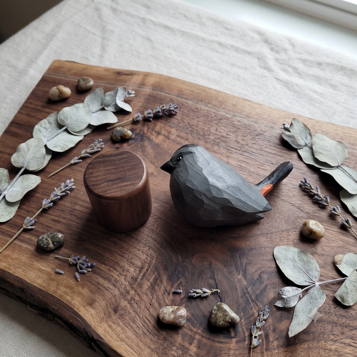 A dark gray carved wooden Gray Catbird figurine with a small orange accent on its tail, displayed on a rustic live-edge wooden board alongside a matching wooden cylinder, surrounded by dried eucalyptus leaves, lavender sprigs, and small stones.