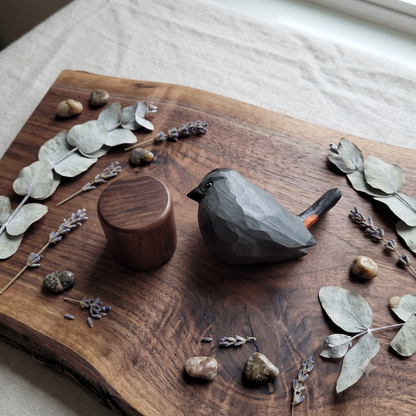 A dark gray carved wooden Gray Catbird figurine with a small orange accent on its tail, displayed on a rustic live-edge wooden board alongside a matching wooden cylinder, surrounded by dried eucalyptus leaves, lavender sprigs, and small stones.