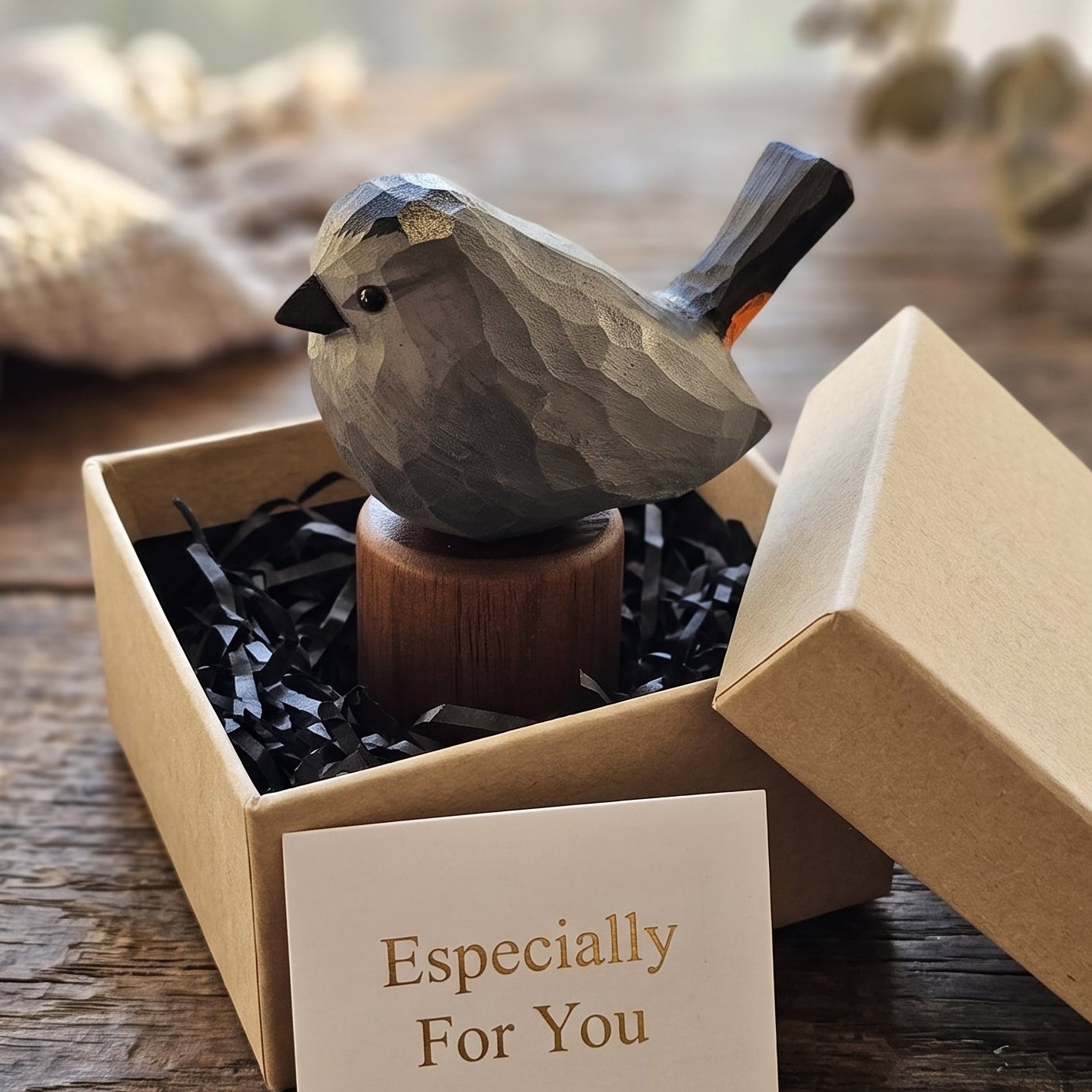 A small carved Gray Catbird figurine in gray and black tones with a dark tail, displayed in an open kraft gift box with black shredded paper and a card that reads 'Especially For You.' The box lid rests beside it on a rustic wooden table, with soft blurred background elements including a cloth and dried stems.