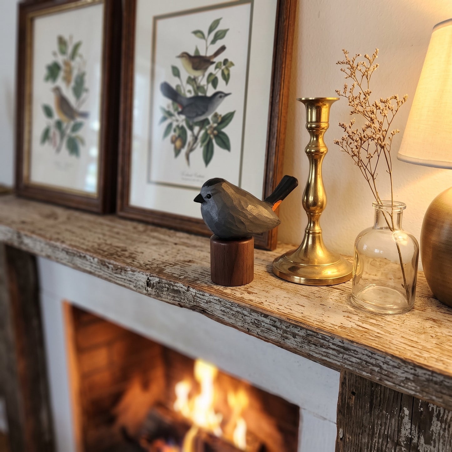 A rustic Gray Catbird figurine on a wooden base displayed on a weathered stone fireplace mantel, styled with framed bird prints, a brass candlestick, a clear glass vase with dried stems, and a lamp in a cozy indoor setting.