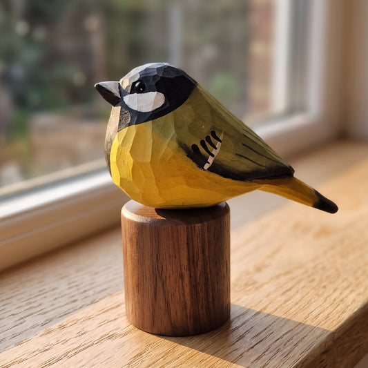 A hand-carved wooden Great Tit figurine, resembling a great tit with yellow, black, and white painted details, perched on a cylindrical wooden base on a wooden windowsill.
