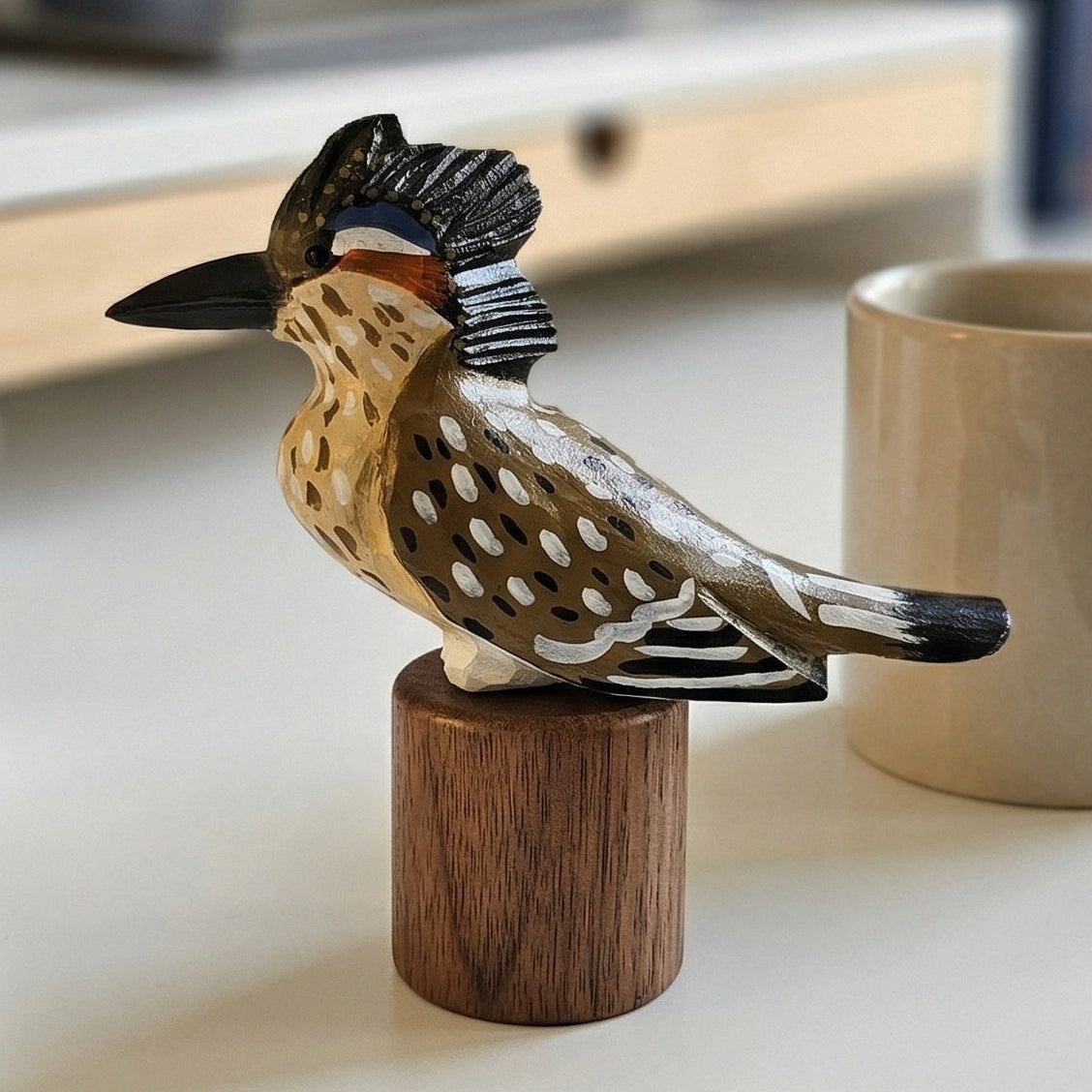 A hand-carved wooden Greater Roadrunner figurine, resembling a kookaburra, perched on a small cylindrical wooden base. The bird features natural wood grain with hand-painted black, brown, white, and orange accents.