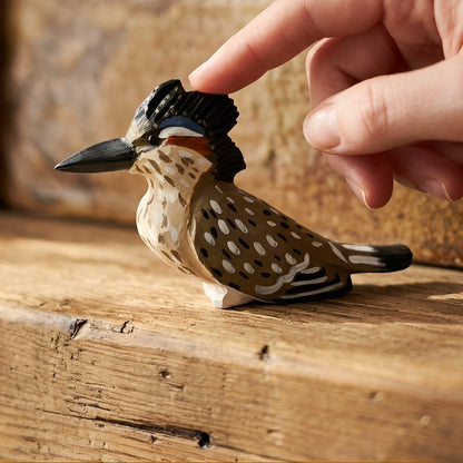 A hand gently touches a small carved wooden Greater Roadrunner figurine with a black crest and spotted brown-and-beige body, placed on a rustic wooden surface.