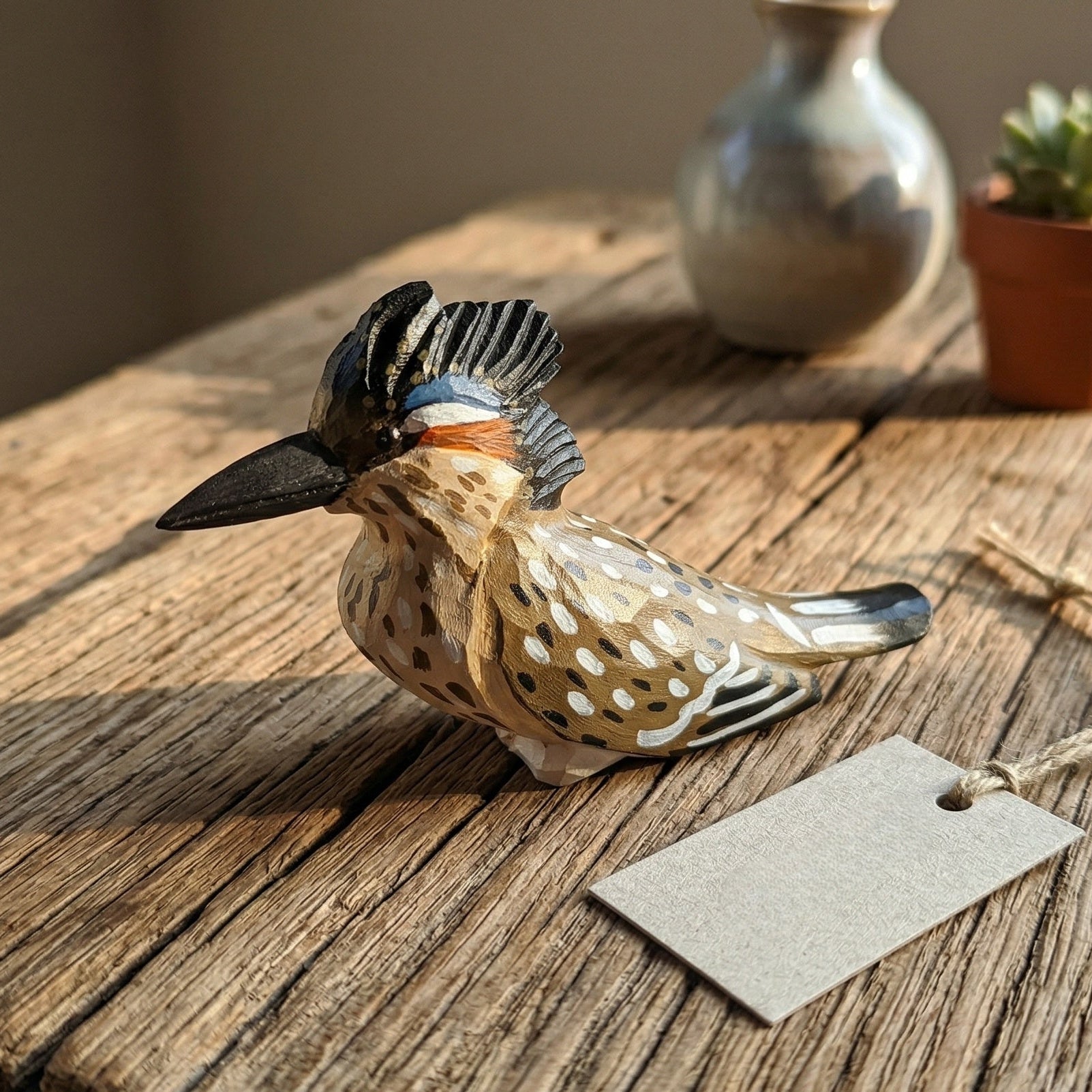 A hand-painted wooden Greater Roadrunner ornament with a black crest and spotted brown-and-white body, resting on a rustic wooden table next to a blank paper tag. In the softly blurred background are a small ceramic vase and a potted succulent.
