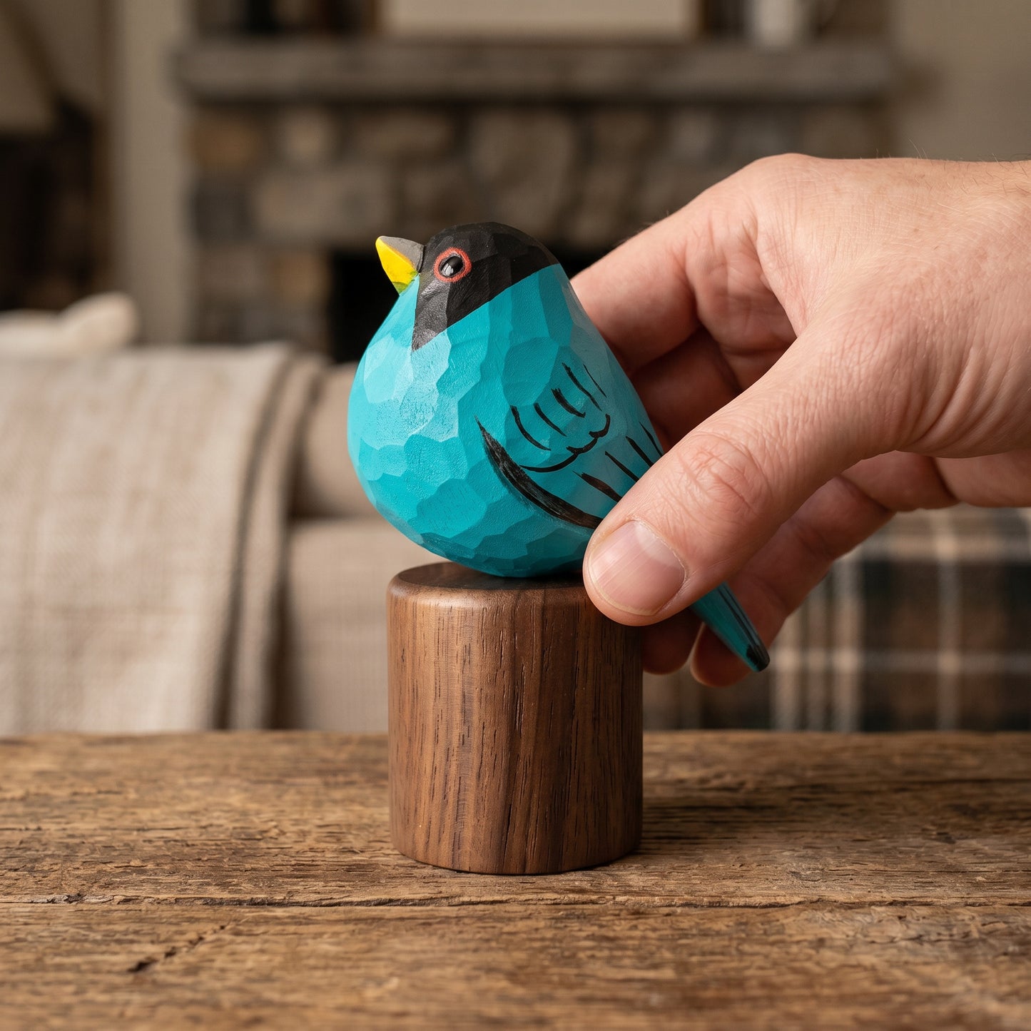 Hand placing a hand-carved turquoise Green Honeycreeper wooden bird figurine onto its cylindrical black walnut base on a rustic wooden table.
