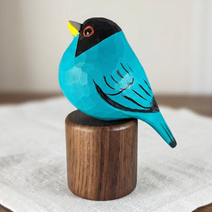 Hand-carved turquoise wooden Green Honeycreeper bird figurine perched on a cylindrical black walnut base on a wooden tabletop.