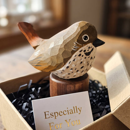 A handcrafted wooden Hermit Thrush  figurine with geometric facets sits in a brown gift box filled with black crinkle paper and a card that reads 'Especially For You'. The background is a softly blurred indoor setting.