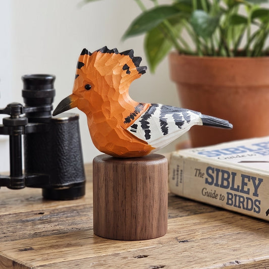 A geometric, low-poly style wooden Hoopoes figurine in orange, black, white, and brown sits on a wooden pedestal on a rustic table. The bird figurine is styled with black binoculars, a book, and a potted plant in the background.