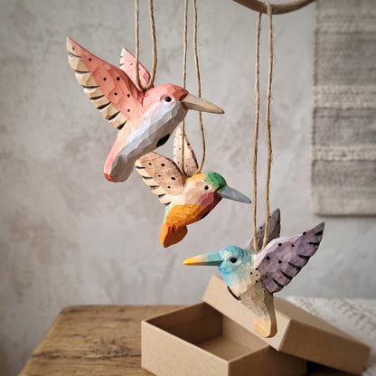 Three hand-carved wooden hummingbird ornaments in pink, orange, and light blue hanging together above a kraft gift box.