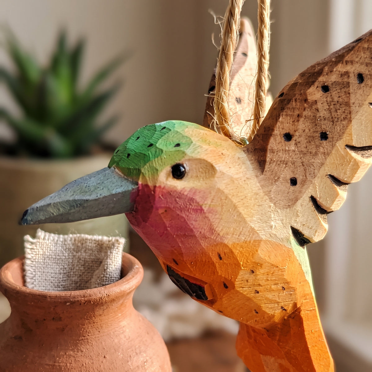 Close-up of a hand-carved wooden hummingbird ornament hanging above a small terracotta pot.