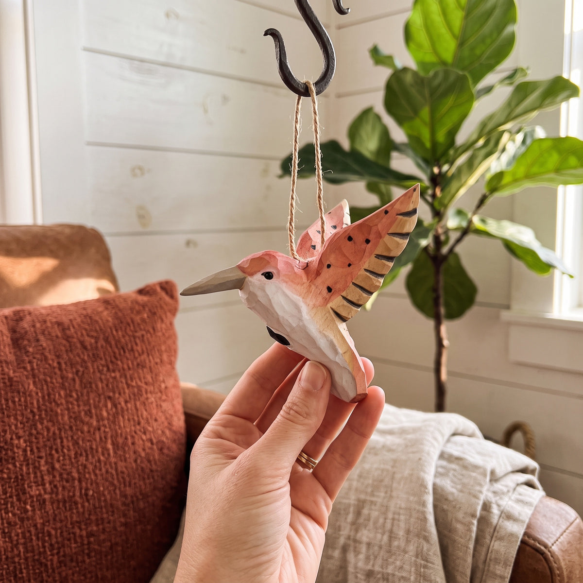 A hand holding a hand-carved pink wooden hummingbird ornament suspended from a black metal hook in a living room.