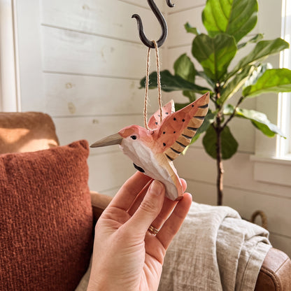 A hand holding a hand-carved pink wooden hummingbird ornament suspended from a black metal hook in a living room.