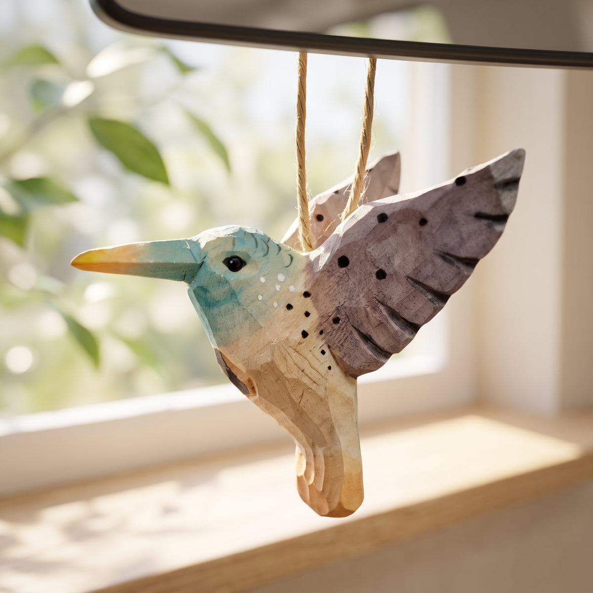 Hand-carved wooden hummingbird ornament with purple and teal accents hanging by a twine string near a sunlit window.