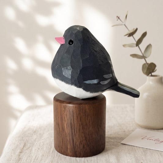 A small Junco figurine with a dark gray and white body and a pink beak, mounted on a round wooden base and styled on a table beside a small ceramic vase with dried leaves.