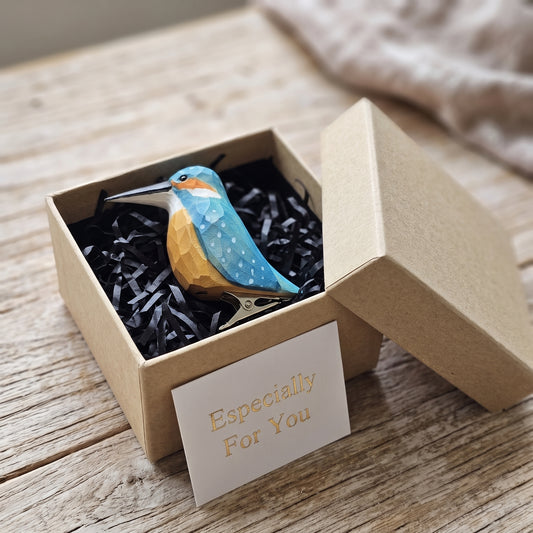 A handcrafted wooden kingfisher clip-on ornament nestled inside an open kraft gift box with black shredded paper, accompanied by a small card reading "Especially For You" on a rustic wooden surface.