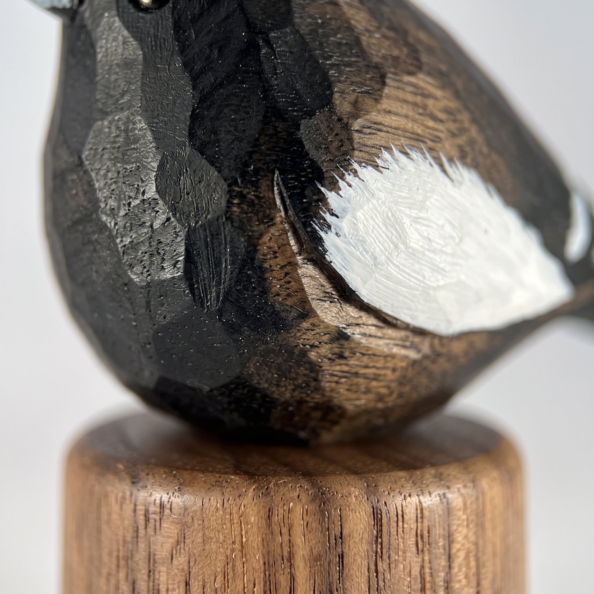 Close-up of a carved wooden Lark bunting figurine with dark black and brown wood grain, a white painted wing, and a round wooden base on a light gray background.